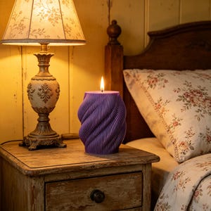 May include: A burning purple candle with a spiral design sits on a wooden bedside table next to a lamp. The candle's flame is lit, creating a warm atmosphere in the bedroom. A bed with floral bedding is in the background.