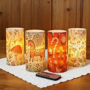 Könnte beinhalten: Vier zylindrische LED-Kerzen mit dekorativen Designs. Eine zeigt einen Pilz, eine andere ein Pferd, eine dritte hat Zuckerstangen und einen Lebkuchenmann, und die letzte hat florale Muster. Eine Fernbedienung liegt vor den Kerzen.