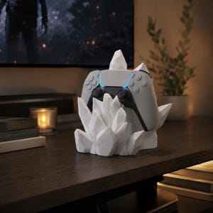 May include: A white, faceted, crystal-shaped gaming controller stand holding a light grey wireless controller. The stand features sharp, angular geometric points that cradle the device, providing a stable base for the controller to rest upon.