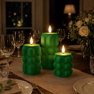 May include: Three cylindrical green candles with a textured, bubble-like surface and vertical ribbing. Each candle features a glowing, flame-shaped LED light at the top. The candles vary in height and are arranged on a table setting with glassware.