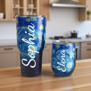 May include: Two insulated tumblers featuring a swirling blue and yellow starry night design. One has the name "Sophia" in white script, the other "Elena". The tumblers are on a wooden surface.