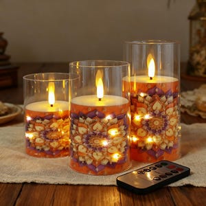 Puede incluir: Juego de tres velas LED sin llama en cilindros de vidrio transparente. Las velas tienen una base naranja con un diseño de mandala púrpura y dorado, envueltas con luces de hadas. Un mando a distancia está en primer plano.