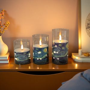 May include: Three battery-operated LED candles in glass cylinders. The candles feature a "Starry Night" design and are lit with a warm, flickering light. The candles are of varying heights and are placed on a wooden surface.