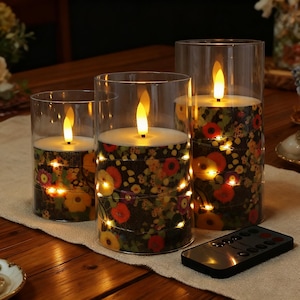 Könnte beinhalten: Drei LED-Kerzen in Glaszylindern. Die Kerzen sind mit einem Blumenmuster und Lichterketten verziert. Eine Fernbedienung befindet sich im Vordergrund. Die Kerzen stehen auf einer beigen Tischdecke. Die Kerzen sind batteriebetrieben.