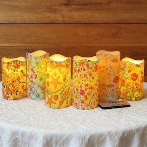 以下が含まれることがあります： 様々なデザインの6つの装飾用LEDキャンドルのセット。花柄、蝶、貝殻、キノコの模様があります。リモコンが付属しています。キャンドルは白い表面に置かれ、背景は木材です。