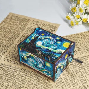 May include: A wooden music box featuring a painted reproduction of Van Gogh's "Starry Night." The box has a crank on the side and is set on a vintage paper background. The artwork depicts a swirling blue night sky with yellow stars.