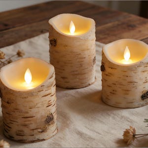 以下が含まれることがあります： ゆらめくLEDの炎が特徴の、白樺風の電池式キャンドル3つ。円筒形で、木の皮のような質感があり、温かみのある光を放っています。クリーム色の布の上に配置され、居心地の良い素朴な雰囲気を醸し出しています。
