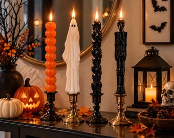May include: Four lit taper candles in candle holders: an orange pumpkin-shaped candle, a white ghost-shaped candle, a black skull-stacked candle, and a black skeleton-shaped candle. The candles are arranged on a dark surface with Halloween decor.
