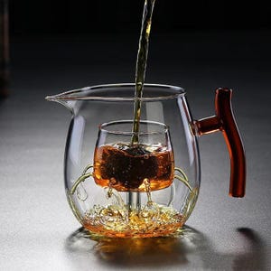 Puede incluir: Una tetera de vidrio transparente con un infusor incorporado y un asa marrón rojizo. Se está vertiendo té en el infusor, que está suspendido dentro de la tetera. El té es de color ámbar.
