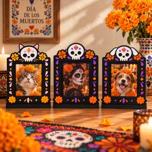 May include: Three black rectangular picture frames with colorful floral and skull motifs. The left frame features cat ears, the center a skull, and the right dog ears. Each frame displays a photograph of a cat, a person, or a dog wearing marigolds.