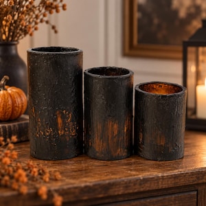 May include: Three cylindrical candle holders of varying heights with a textured black exterior finish and visible orange-brown undertones. The hollow, open-top vessels feature a rugged, bark-like surface appearance, arranged in a row on a flat surface.