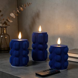 May include: Three navy blue decorative candles with a textured, bubble-like design. Each candle has a flickering, warm-toned LED flame. A black remote control is in the foreground. The candles are on a grey surface.