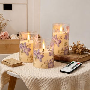 May include: Three flameless candles in glass holders, decorated with purple floral patterns. The candles are lit, and a remote control sits nearby. The candles are on a small, round table covered with a white, textured cloth.