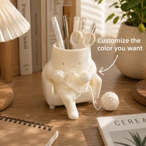 May include: A white desk organizer shaped like a character sitting in a chair and knitting with yarn. The character has a smiling face and holds pens and scissors. Text on the image reads: Customize the color you want, with an arrow pointing to the item.