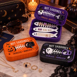 May include: Three rectangular metal tins labeled NOT SARDINES SKINLESS BONELESS. One purple tin is open, revealing white fish-shaped jewelry organizers holding rings, earrings, and necklaces. Two closed tins, one orange with a pumpkin and one black with a ghost.