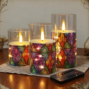Op de afbeelding: Drie LED-kaarsen in glazen cilinders. De kaarsen hebben een kleurrijk glas-in-loodontwerp met rode, oranje, gele, groene en paarse tinten. Elke kaars heeft een flikkerend vlameffect en is omwikkeld met lichtslingers. Een afstandsbediening staat op de voorgrond.