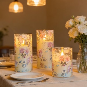 Op de afbeelding: Drie decoratieve kaarsen in glazen houders, met een bloemenrozenontwerp in roze en paarse tinten. De brandende kaarsen creëren een warme sfeer. Ze staan op een tafel met een wit tafelkleed.