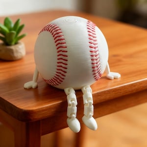 Puede incluir: Una pelota de béisbol blanca con costuras rojas y brazos y piernas blancos adjuntos, sentada sobre una superficie de madera. La pelota de béisbol tiene un diseño caprichoso, lo que le da una apariencia lúdica. Es un artículo decorativo único.