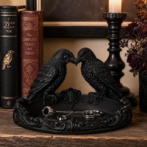 May include: A black decorative tray featuring two sculpted ravens facing each other on the rim. The tray has an ornate, raised border with intricate scrollwork details. Inside the tray, a silver chain necklace and two rings are resting.