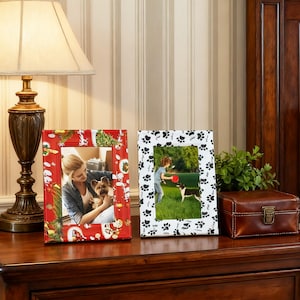 May include: Two decorative picture frames on a wooden surface. One frame is red with a festive design, the other is white with black paw prints. The frames hold photos of a woman with a dog and a child playing with dogs.