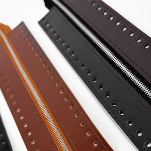 May include: Three leather zipper strips in black, brown, and tan, each with a silver zipper and evenly spaced holes along the edges. The zippers are open, revealing the silver teeth. The strips are arranged diagonally against a white background.