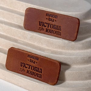 Op de afbeelding: Twee rechthoekige bruine leren labels met de tekst "MADE -by- VICTORIA IONOVA" in donkerbruin geëmbosseerd. De labels liggen op een lichtbeige, trapsgewijs, gebogen oppervlak.