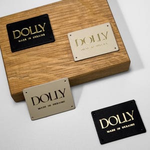 Puede incluir: Cuatro etiquetas rectangulares con la palabra "DOLLY" en letras doradas. Dos etiquetas son negras y dos son beige. El texto "MADE IN UKRAINE" está debajo del nombre de la marca. Las etiquetas se muestran en un bloque de madera.