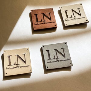 May include: Four square leather tags in various colors: brown, beige, and gray. Each tag features the letters "LN" and the words "knitwears & accessories" engraved in a dark color. The tags have small holes in each corner.