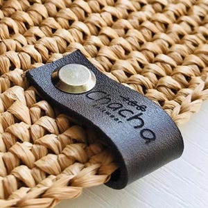 Puede incluir: Primer plano de un sombrero de paja tejido con una banda azul estampada. El sombrero presenta una etiqueta de cuero marrón oscuro con la marca "Chacha" y un botón plateado. El tejido del sombrero es de color beige natural.