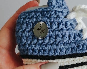 May include: A handmade, blue and white crochet baby sneaker with white laces and a black stripe. The sneaker has a small metal tag with initials. The shoe is held in a hand.