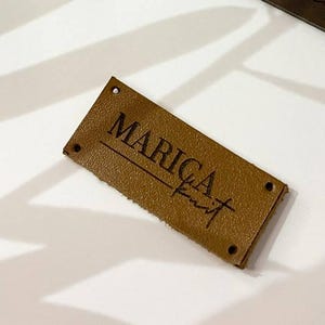 Puede incluir: Cuatro etiquetas rectangulares de cuero con el texto "MARICA knit" en escritura a mano. Las etiquetas son en tonos marrones y están dispuestas sobre una superficie blanca con sombras de hojas. Cada etiqueta tiene dos pequeños agujeros en cada extremo.