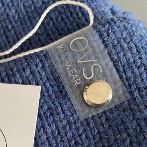May include: A close-up shot of a blue knitted hat with a tag attached. The tag displays "OVS Knitwear handmade" with a floral design and the Instagram handle "@ovs_kw". A small, clear tag with "OVS Knitwear" is also attached.