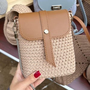 Op de afbeelding: Een beige gehaakte crossbodytas met een bruine leren flap en riem. De tas heeft een zilveren kettingriem en een knoopsluiting. Andere gehaakte tassen in verschillende kleuren staan op de achtergrond.