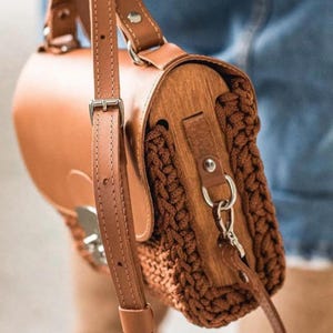 Può includere: Una borsa in pelle e legno marrone con un pannello laterale marrone intrecciato. La borsa ha una lunga tracolla regolabile e una chiusura color argento. La borsa è tenuta da una persona che indossa jeans blu e stivali marroni.