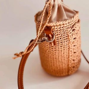 Pode incluir: Uma bolsa tipo balde cilíndrica feita de palha trançada na cor castanho com um fecho de cordão de tecido bege na parte superior. A bolsa possui uma alça de ombro de couro marrom liso presa com ferragens de metal prateado e um cordão trançado correspondente.
