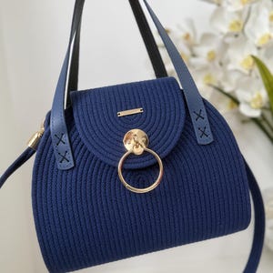 May include: A navy blue handbag crafted from coiled cord material, featuring a rounded flap closure with a prominent gold-toned circular metal ring accent. The bag includes two blue leather-like straps with decorative X-stitching at the base.