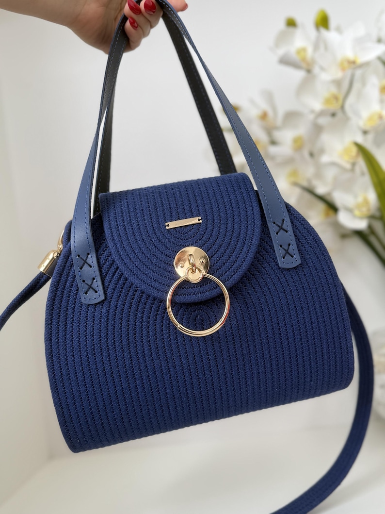 May include: A navy blue handbag crafted from coiled cord material, featuring a rounded flap closure with a prominent gold-toned circular metal ring accent. The bag includes two blue leather-like straps with decorative X-stitching at the base.