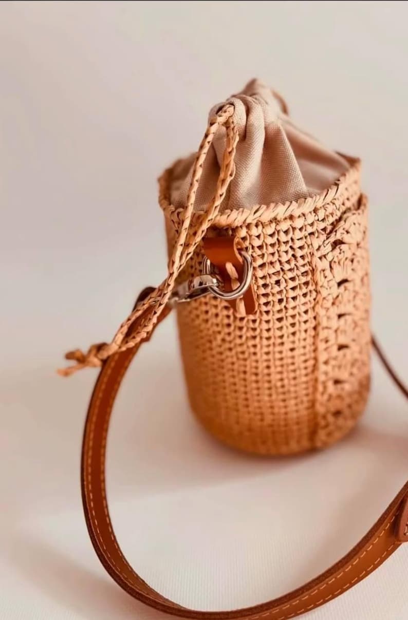 May include: A cylindrical bucket bag crafted from woven tan straw with a beige fabric drawstring closure at the top. The bag features a smooth brown leather shoulder strap attached with silver-toned metal hardware and matching braided cord.