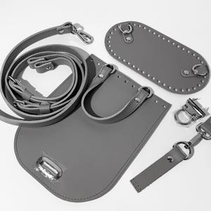 May include: A grey leather bag making kit featuring a rounded base panel with pre-punched holes, a main body piece with attached handles, a long adjustable shoulder strap, and silver-toned metal hardware including D-rings and a clasp closure.