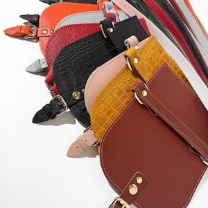 May include: A collection of colorful leather bags and straps in various shades, including brown, black, orange, and yellow. The bags have a half-moon shape with gold-colored hardware and adjustable straps. The leather textures vary, with some bags featuring a crocodile pattern.