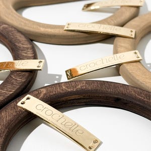 Puede incluir: Asas de bolso de madera en varios tonos de marrón. Cada asa tiene un diseño curvo y se combina con una etiqueta metálica dorada que dice "crochelle". Las etiquetas también tienen texto en otro idioma.