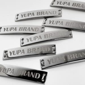 May include: A collection of shiny, silver metal tags with the text "YUPA BRAND" engraved on them. Each tag has rounded edges and two small holes on each end. The tags are scattered on a white surface.