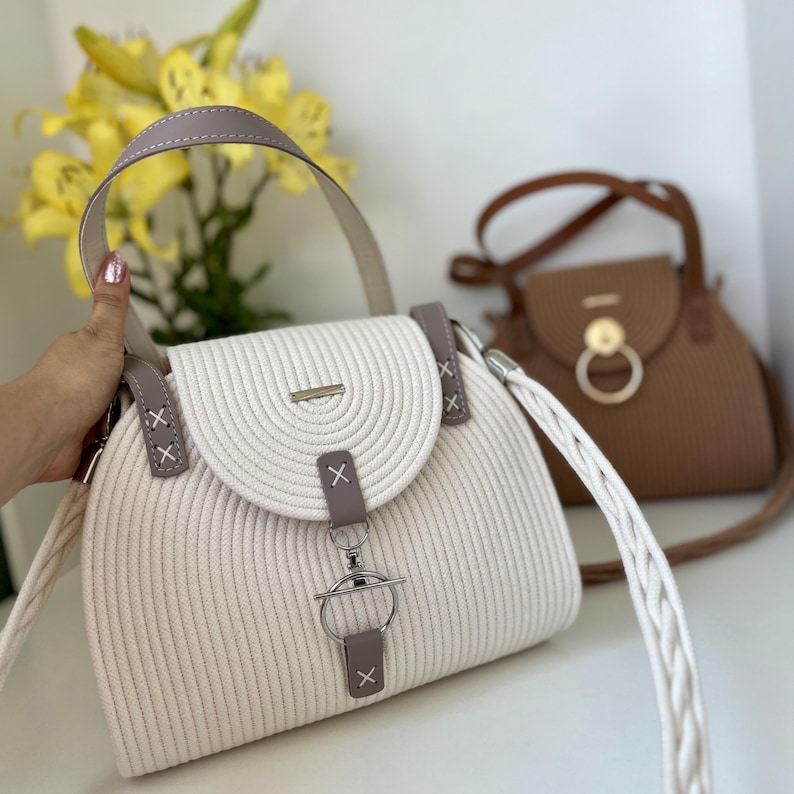 May include: A cream-colored handbag with a ribbed texture, featuring a rounded flap, a top handle, and a braided shoulder strap. The bag is accented with grey leather-like tabs, silver-toned hardware, and a circular metal ring closure.