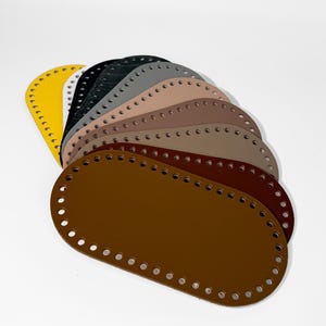 Könnte beinhalten: Eine Sammlung ovaler Leder-Taschenböden in verschiedenen Farbtönen, darunter Gelb, Schwarz, Grau und Braun. Jeder Boden hat gleichmäßig verteilte Löcher am Umfang zum Befestigen an einem Taschenkorpus. Die Böden sind fächerförmig angeordnet.