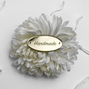 May include: An oval-shaped gold-colored mirror tag with the word Handmade engraved in a cursive script font. The tag features two small holes on the left and right sides and is positioned on top of a large, white, multi-petaled flower.