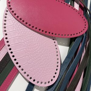 Könnte beinhalten: Eine Sammlung ovaler Lederböden und langer, dünner Lederriemen in verschiedenen Farben wie Pink, Hellrosa, Blau, Grün und Braun. Die ovalen Böden haben vorgestanzte Löcher an den Rändern und die Riemen weisen eine strukturierte Oberfläche auf.