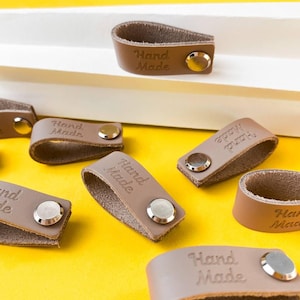 May include: A collection of brown leather labels folded into loops and secured with silver-toned metal rivets. Each label features the embossed text Hand Made. The leather has a smooth finish on the exterior and a textured surface on the interior.