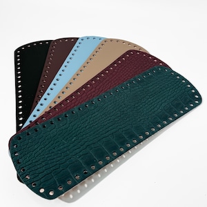 May include: A collection of colorful, rectangular bag bases in various shades, including black, brown, blue, tan, burgundy, and dark green. Each base has a series of evenly spaced holes along the edges for attaching to a bag. The dark green base has a crocodile skin texture.