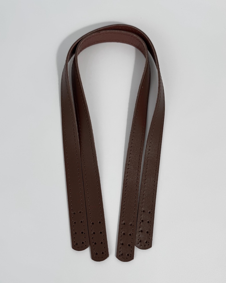 May include: A pair of dark brown bag handles made of smooth material with a matte finish. Each strap features a long, slender design with visible stitching along the edges and four pre-punched holes at each end for secure attachment to a bag.