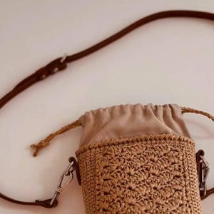 May include: A small cylindrical crossbody bag featuring a tan crocheted raffia body with a textured shell-like pattern. The bag has a beige fabric drawstring closure at the top, a thin brown leather strap with silver metal hardware, and side clips.